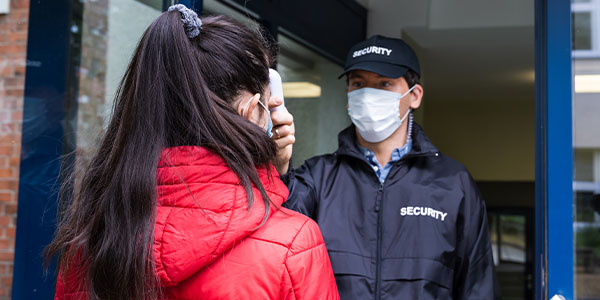 Young male security guard with surgical mask on, checking the temperature of a female in a red coat